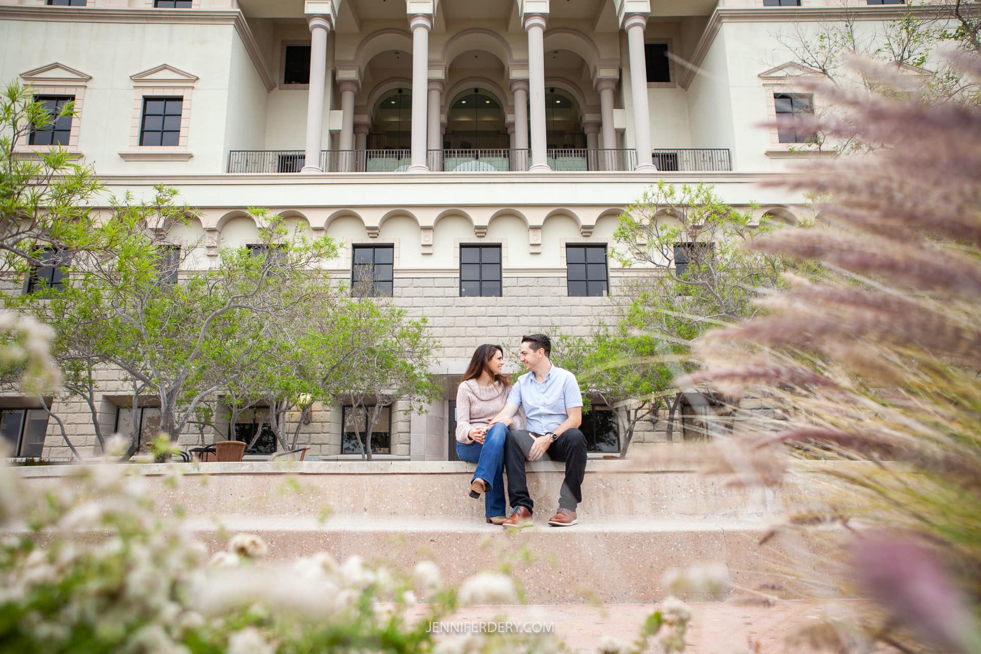 A couple sits on a stone ledge in front of a large, elegant building with multiple arches and balconies at USD campus. Surrounding them are green plants and grasses, creating a serene and picturesque setting. The man looks at the woman while she gazes in front.