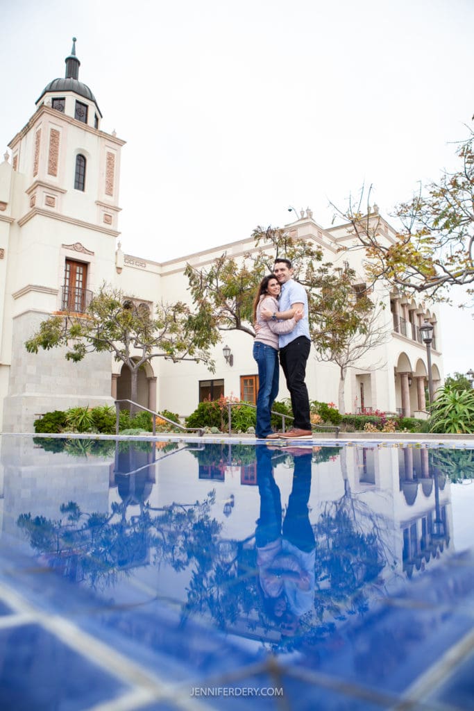An engagement session couple stands embracing in front of a historic building at USD with a tower. They are reflected in the blue-tiled surface of a shallow pool in the foreground. Surrounding the building are trees and shrubs. The sky appears overcast.