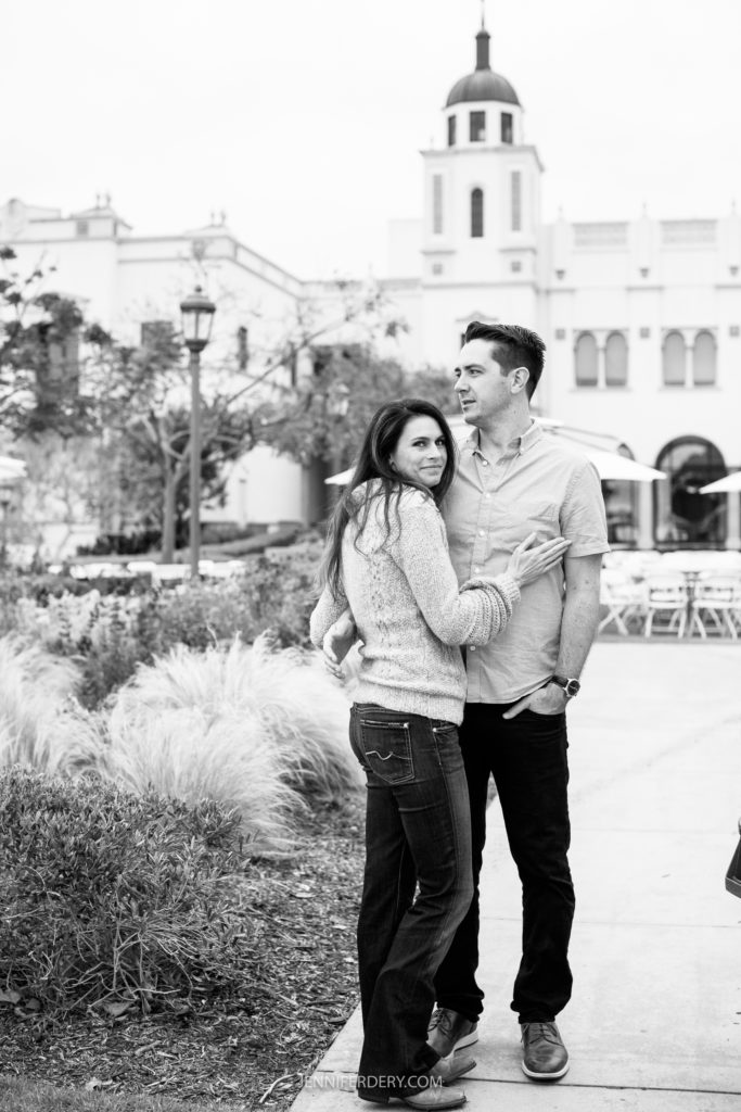 A engagement session couple stands outdoors in front of a building with a bell tower at the USD campus. The woman, wearing a sweater and jeans, looks directly at the camera while the man, in a button-up shirt and jeans, gazes into the distance. They stand closely together, with her arm wrapped around his waist.