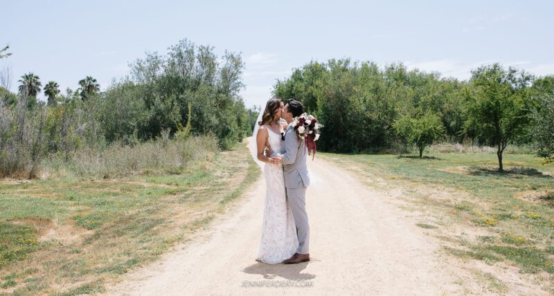 A bride and groom share a kiss on a dirt path at rancho guajome surrounded by greenery. The bride, in a white gown and veil, holds a bouquet of flowers. The groom, in a light grey suit, embraces her. Trees and bushes line the path under a clear sky.