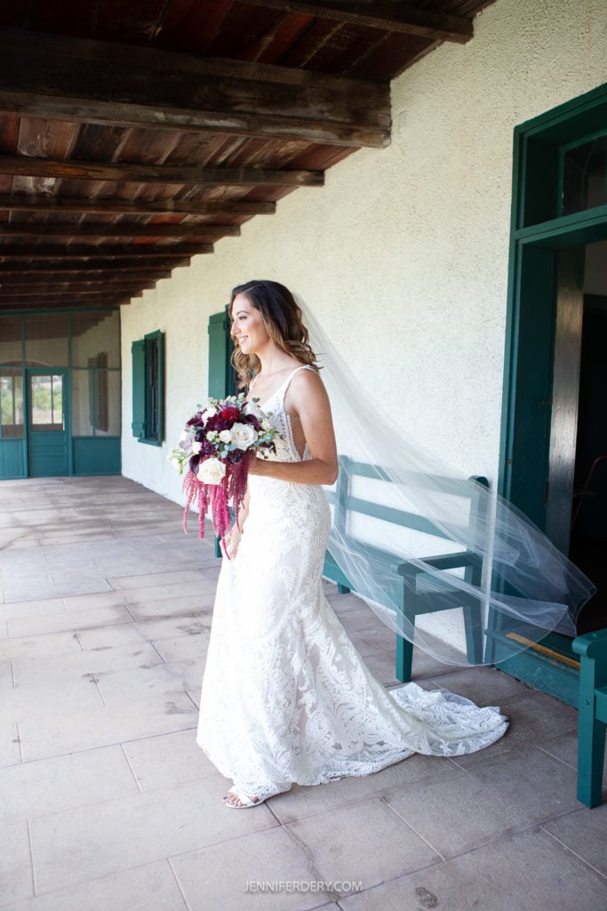 at Rancho Guajome Adobe A bride stands on a porch in a sleeveless lace wedding gown, holding a bouquet with white, burgundy, and green flowers. A long veil flows behind her. The porch has green window frames and doors, with a wooden roof, giving a rustic charm to the scene.