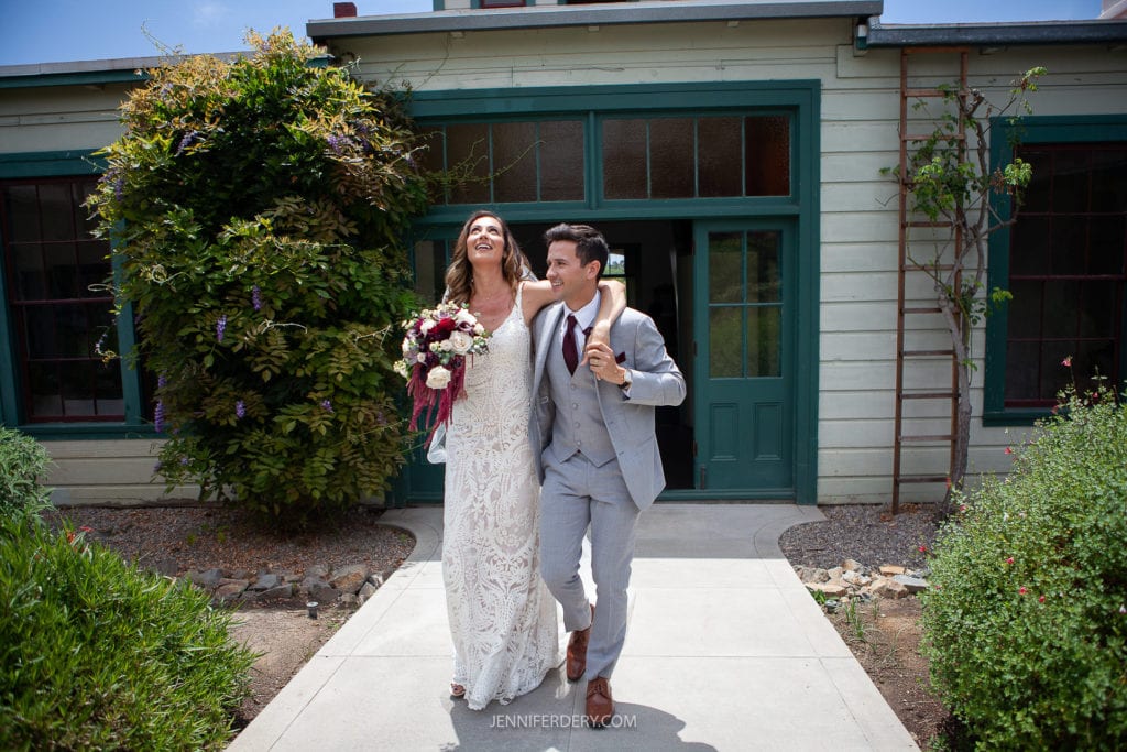 A bride in a white lace gown and a groom in a light gray suit walk joyfully arm in arm down a path at Rancho Guajome Adobe. The bride holds a bouquet of flowers and both appear to be laughing. They are in front of a green and beige building surrounded by greenery.