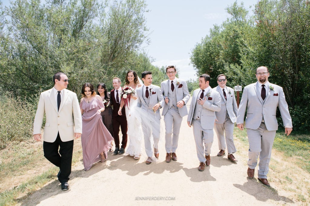 A wedding party walks down a path surrounded by greenery. The bride, in a white gown, and the groom, in a light grey suit, are at the center. Bridesmaids in lavender dresses and groomsmen in matching grey suits with burgundy ties walk alongside them, smiling.
