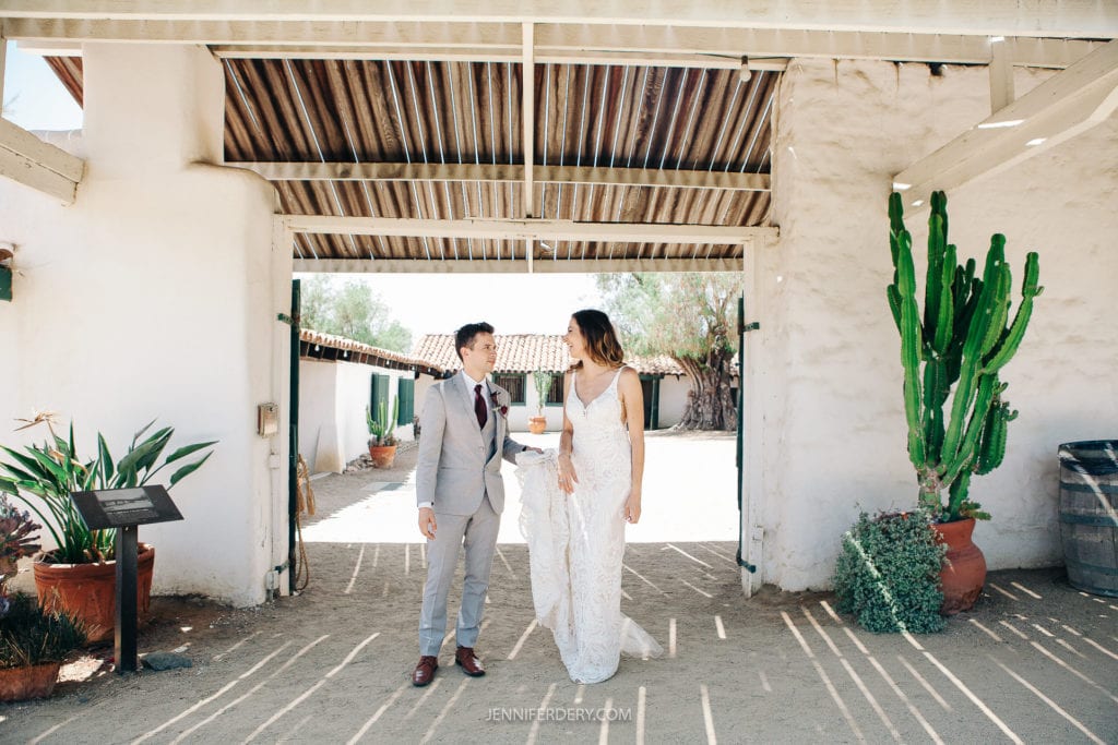 A couple is standing and holding hands under a covered archway at Rancho Guajome Adobe. The groom is wearing a grey suit and the bride is in a lacey white gown. Potted plants, including a large cactus, are visible nearby. Sunlight casts shadows on the sandy ground.