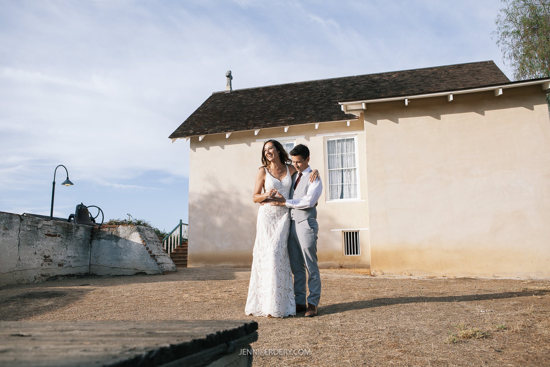 at Rancho Guajome Adobe A couple stands outside in front of a beige building with a pitched roof under a bright blue sky. The woman is wearing a white lace dress, and the man is in gray pants and a white shirt. They are embracing and smiling. The ground is dry and barren.