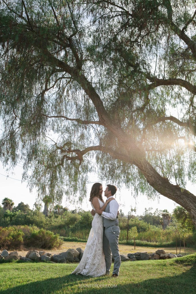 A couple in wedding attire share an intimate moment under a large, leafy tree with sunlight streaming through the branches. They're standing in a grassy area at Rancho Guajome Adobe surrounded by scenic greenery and rocks, with a soft, romantic atmosphere.
