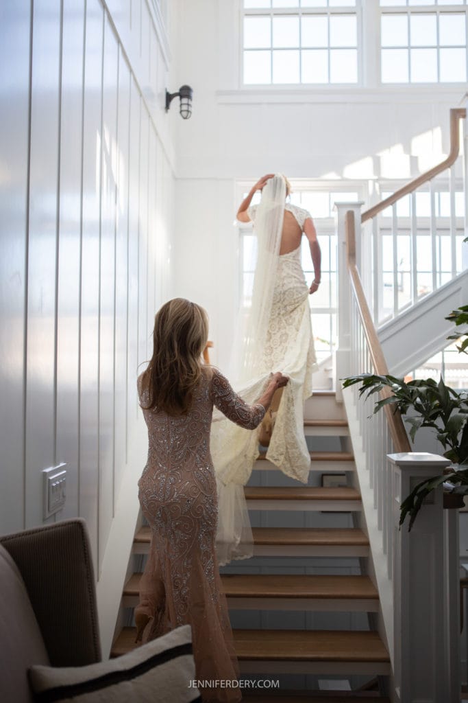 A bride, dressed in a long lace gown, walks up a well-lit staircase while holding her veil, followed by another woman in an embellished dress who assists with the train of the bridal gown. Sunlight streams in through large windows above, illuminating the scene.