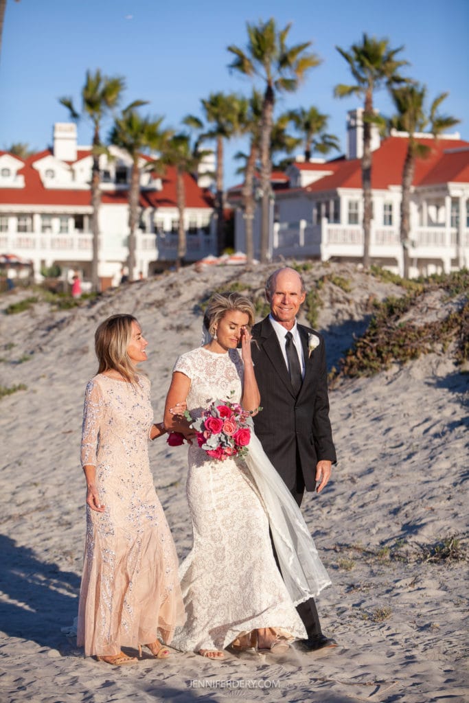 A bride in a white lace dress holds a bouquet of red and pink flowers while walking on a sandy beach with a man in a black suit and a woman in a pale pink dress. In the background are palm trees and white buildings with red roofs.