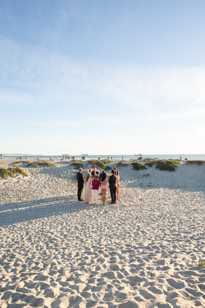 A group of people, dressed formally, stand together on a sandy beach under a clear blue sky. They appear to be part of a special occasion, possibly a wedding or formal gathering. In the background, others walk along the shoreline and enjoy the beach scenery.
