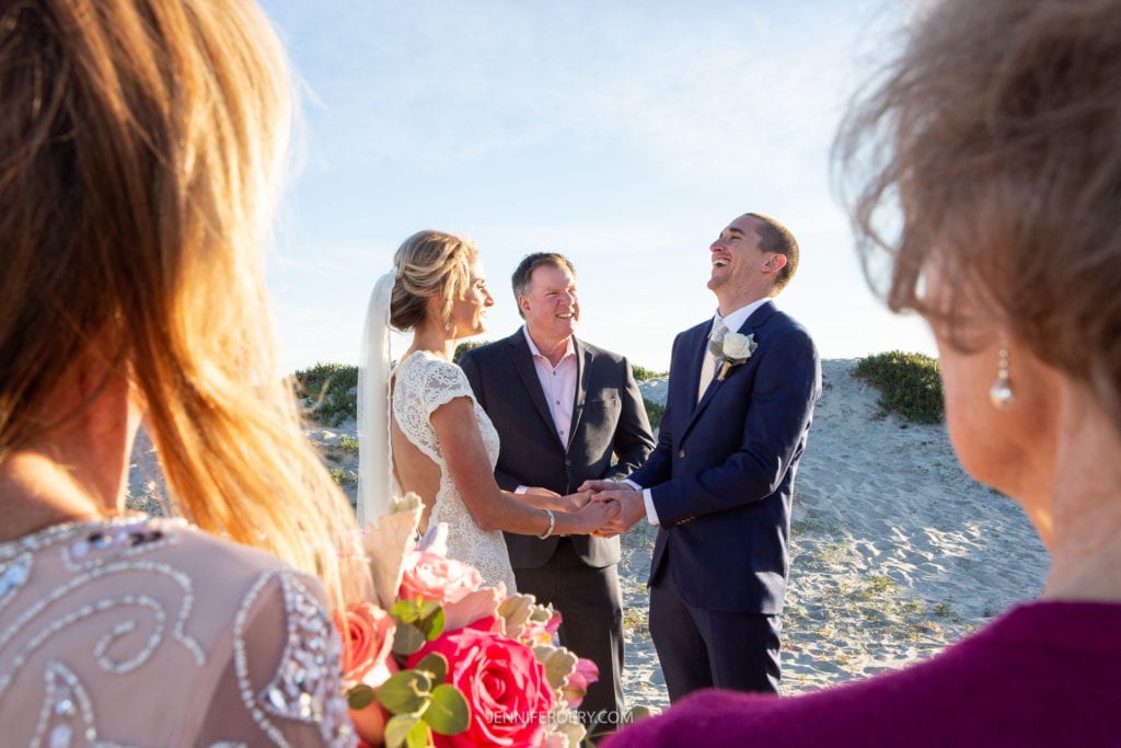A bride and groom hold hands and laugh with a suited officiant on a sandy beach during their wedding ceremony. The bride wears a white dress and veil, while the groom dons a blue suit with a white rose boutonniere. Two guests partially visible in the foreground hold flowers.