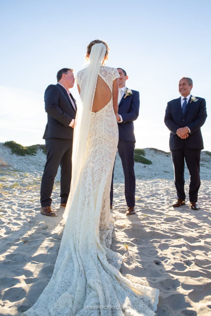 A bride in a white lace gown with a long train stands facing the groom and officiant, holding hands with the groom during an outdoor beach wedding ceremony. Two men in suits stand beside the couple, all smiling. The backdrop includes sand dunes and a clear sky.