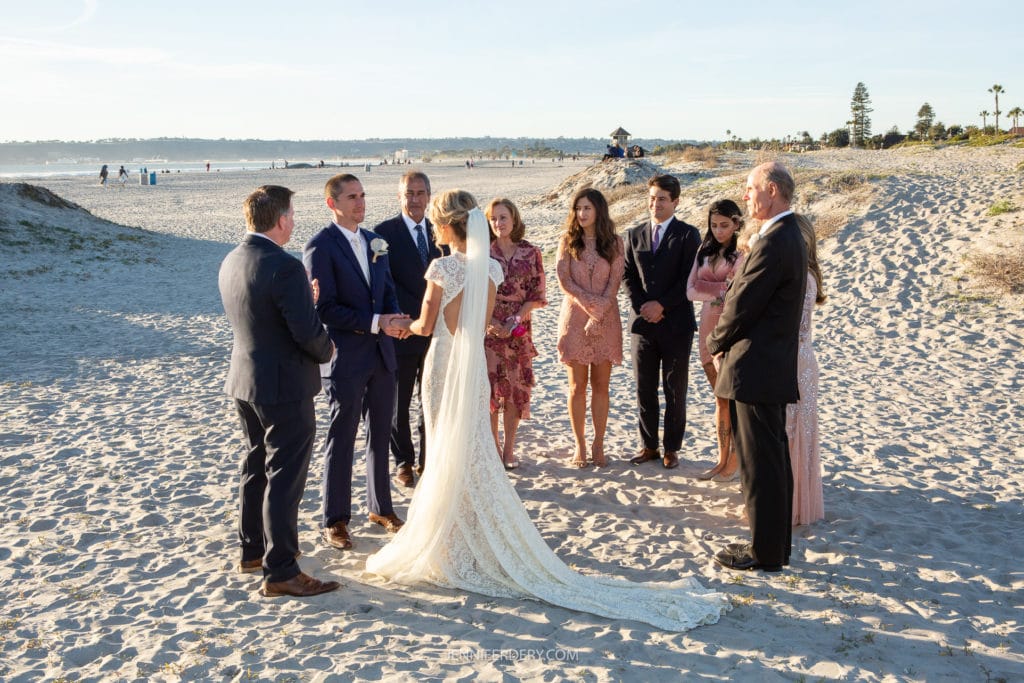 A wedding ceremony on a sandy beach in coronado with a bride in a long white lace dress and a groom in a dark suit. The couple faces each other, holding hands, surrounded by family and friends dressed in formal attire. The ocean and a clear sky are visible in the background.