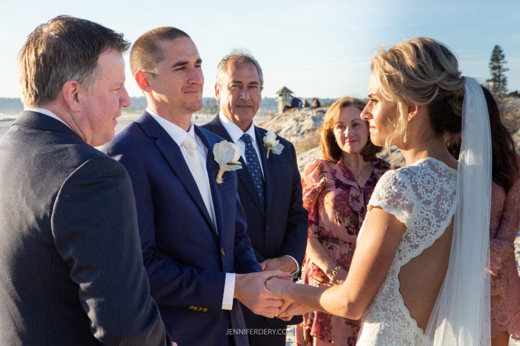 A bride and groom stand facing each other and holding hands in an outdoor wedding ceremony by the beach. The groom has a shaved head and wears a dark suit with a white boutonniere, while the bride wears a lace dress and veil. Several guests stand nearby, smiling.
