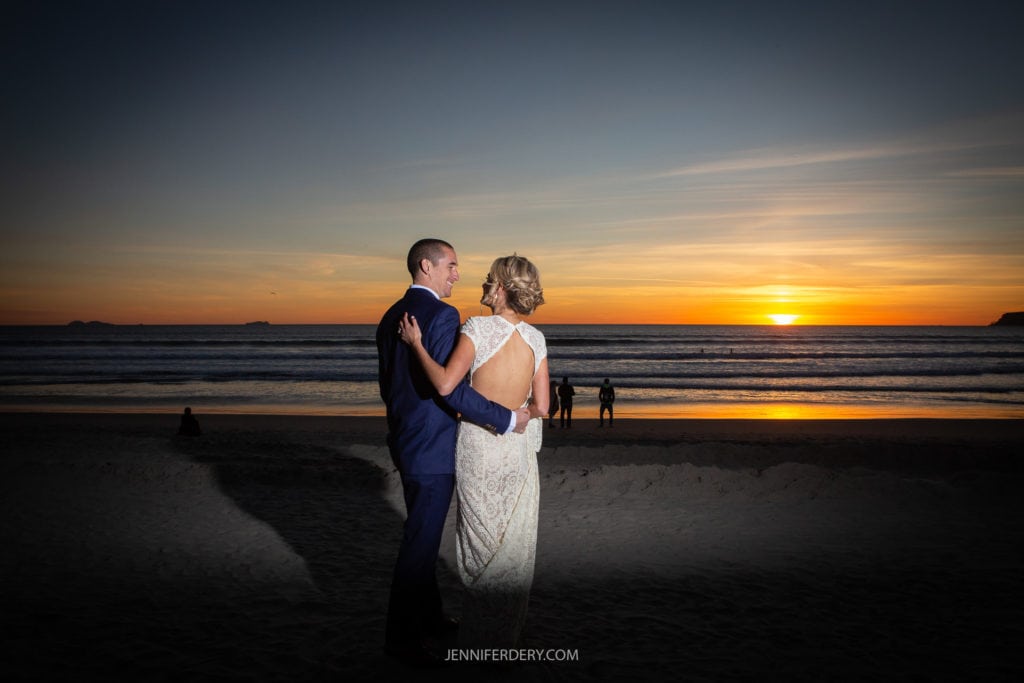 A couple in elegant attire stands close together on a beach, facing the ocean during sunset in coronado. The woman in a white dress rests her hand on the man's shoulder, while he wears a dark suit. The sky is beautifully lit with shades of orange and purple, and a few people can be seen in the distance.