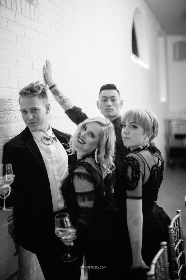 A black-and-white photo shows two men and two women dressed in formal attire, posing and smiling at the camera. They each hold a glass of wine. The setting appears to be an indoor event with white brick walls and large windows.