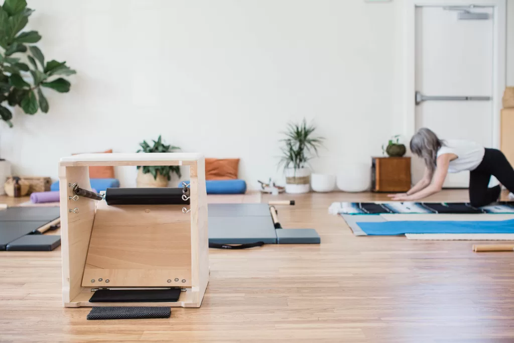 A serene Pilates studio captured by a San Diego branding photographer showcases a wooden exercise apparatus in the foreground and a person with gray hair in athletic wear stretching on a mat in the background. The studio features wooden floors, minimalistic decor, and potted plants.