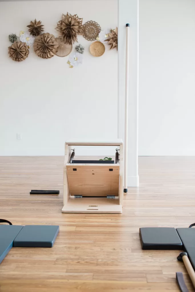 A minimalist Pilates studio in San Diego features a wooden reformer machine at its center, surrounded by light blue mats on a polished wooden floor. The white walls, adorned with decorative paper flowers and intricate circular designs in neutral tones, make it an ideal setting for a San Diego branding photographer.