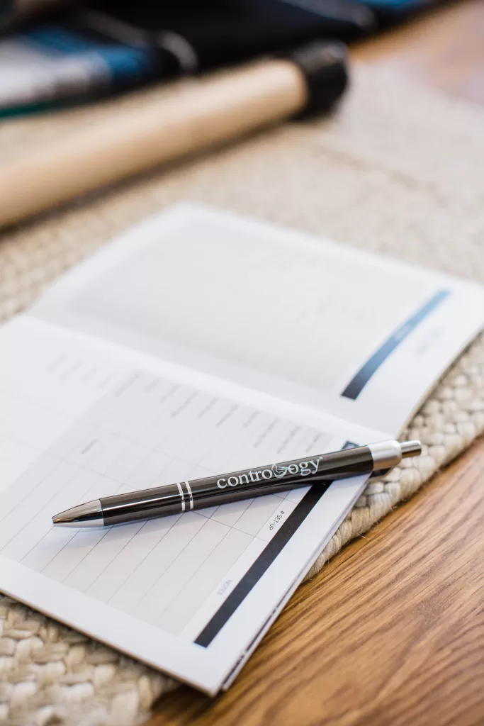 A black pen with the word "contrology" printed on it rests on an open notebook with lined pages. The notebook is placed on a woven fabric, and there's a wooden surface and another object, possibly a stick, in the background—captured flawlessly by a San Diego branding photographer.