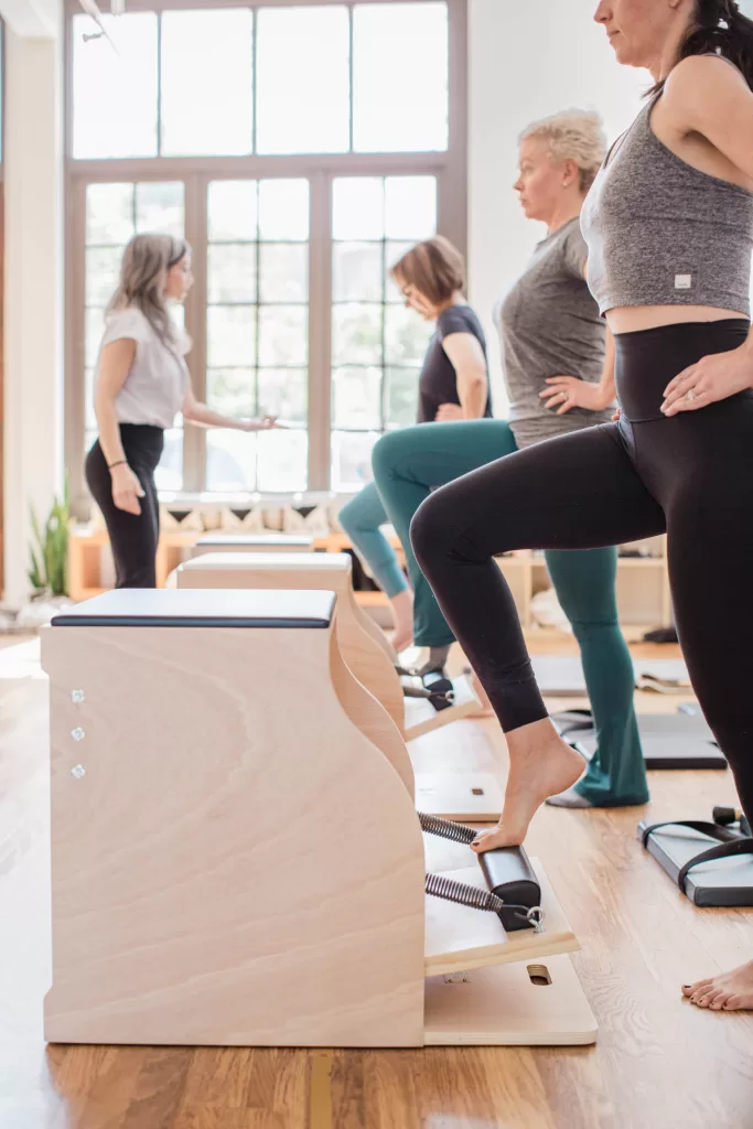 Four women engage in a Pilates class in a bright, sunlit studio captured by a San Diego Branding Photographer. Three women perform leg exercises on wooden Pilates chairs while one instructor observes and guides them. The room features large windows, providing ample natural light, and wooden floors.