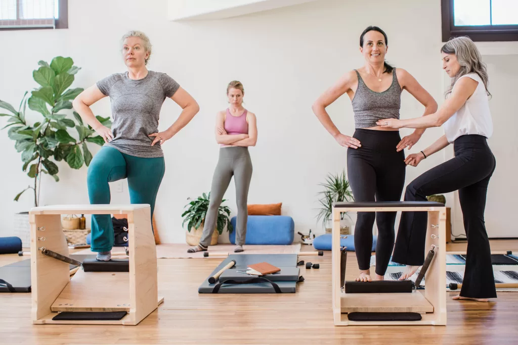 Four women are in a fitness studio. Two women are stepping on Pilates chairs, with classical pilates instructor Nicole Martin assisting one of them. Another woman stands in the background observing. Exercise mats and plants decorate the well-lit studio captured by a San Diego Branding Photographer.