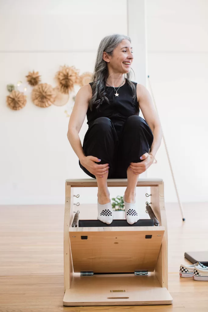 A woman with long gray hair, wearing a black sleeveless top and black pants, sits cross-legged on a wooden Pilates chair. She is smiling and looking to her right. Captured by a San Diego branding photographer, the scene includes wall decorations and a wooden floor in the background.