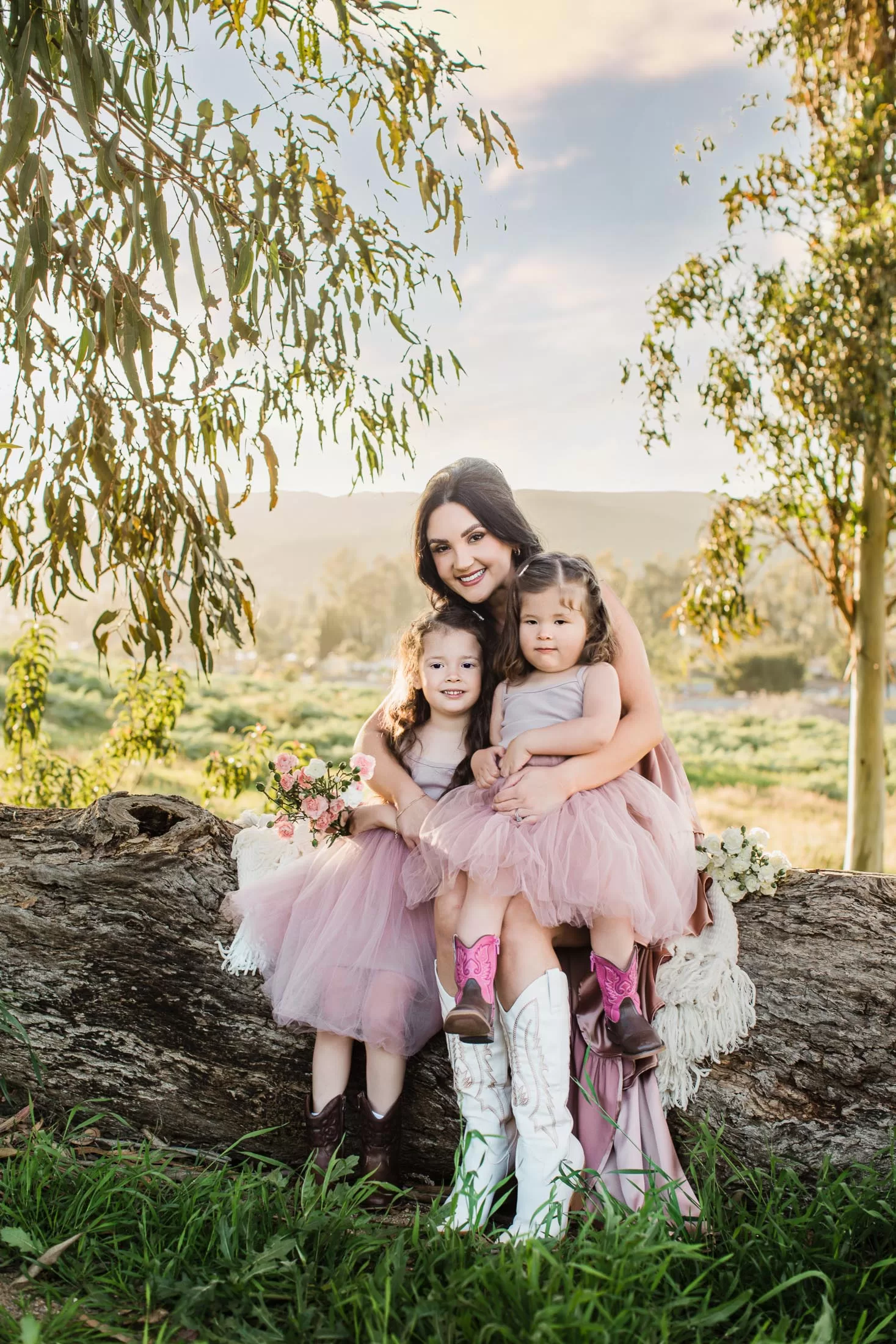 mother's day session showing A woman sits on a large fallen tree trunk outdoors, holding two young girls on either side of her. All three are wearing matching pink dresses, and the girls have cowboy boots. The background features greenery and trees under a bright, partly cloudy sky.