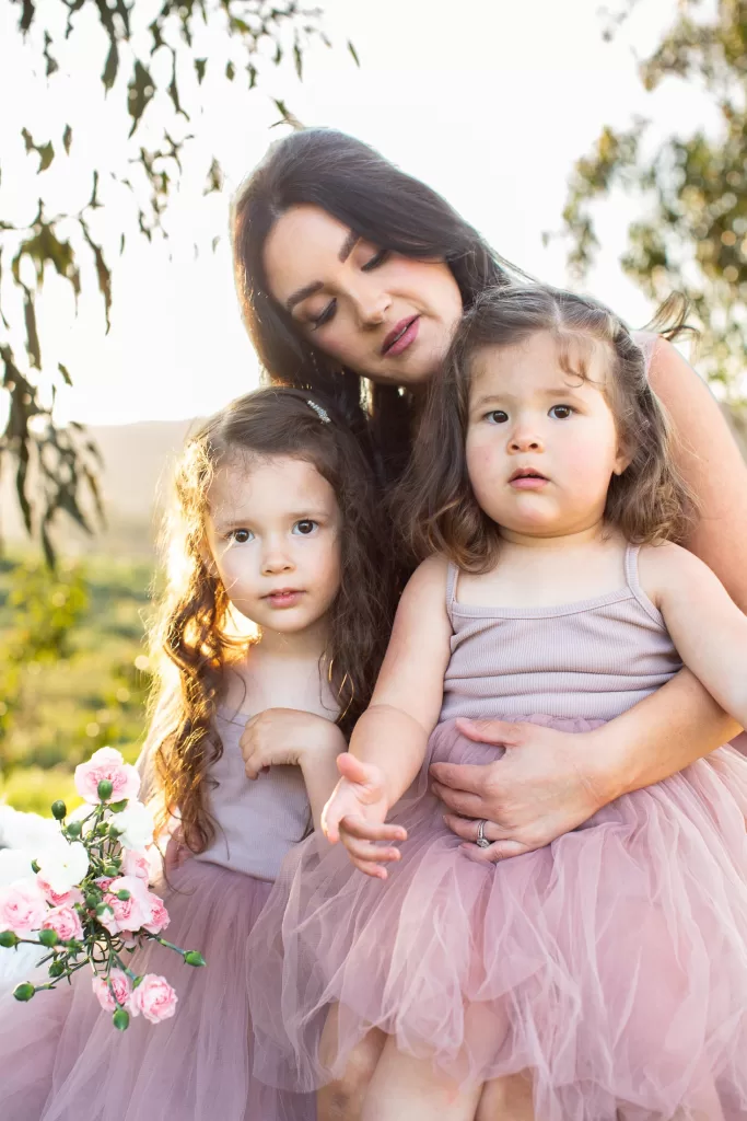 mother's day session showing A woman with long dark hair embraces two little girls wearing matching pink dresses. They are outdoors in a sunny, green setting with trees and pink flowers visible. The children have curly hair and are looking towards the camera, while the woman looks down at them.