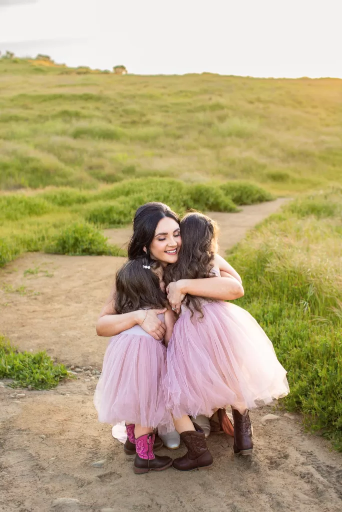 mother's day session showing a woman with long dark hair embraces two young girls in pink dresses on a dirt path surrounded by green fields. The woman is squatting down, smiling warmly, and the girls' backs are to the camera. The scene is illuminated by soft sunlight.