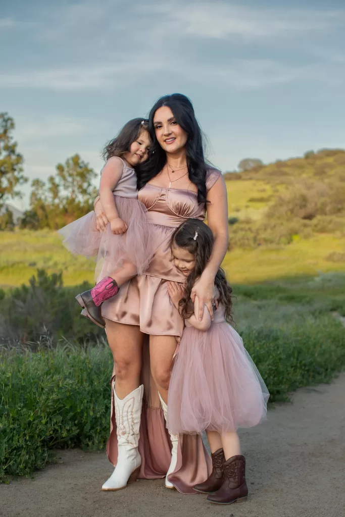 mother's day session showing a woman with long dark hair, wearing a short satin dress and white cowboy boots, stands outside in a grassy field. She holds a young girl in a pink dress and brown boots, while another young girl in a similar pink dress hugs her leg. The sky is cloudy.