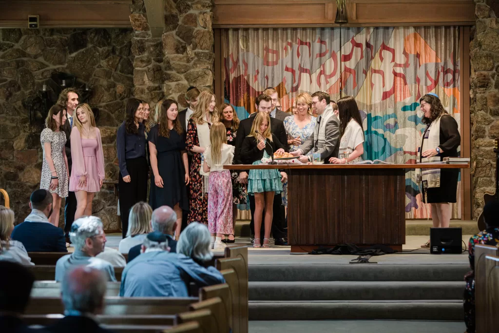 A group of people stand around a table on a stage in a synagogue during a Palo Alto Bat Mitzvah. The backdrop displays Hebrew text with colorful designs. The congregation is seated in pews, attentively watching. An individual at the podium appears to be leading the group in an activity.