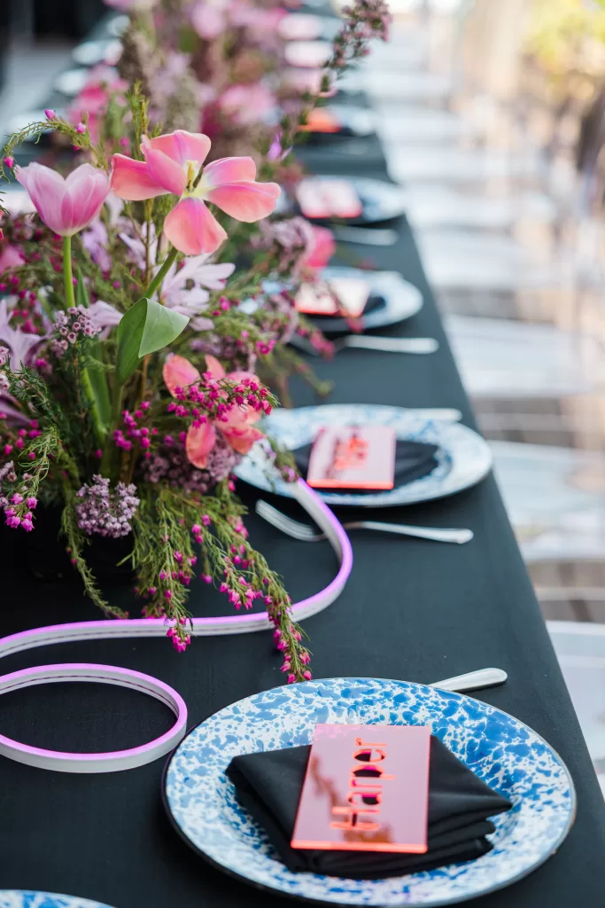 A beautifully set table for a Palo Alto Bat Mitzvah features a black tablecloth, white and blue patterned plates, black napkins, and bright pink name cards. Centerpieces of pink tulips and purple flowers add a vibrant touch to the elegant, modern table setting.