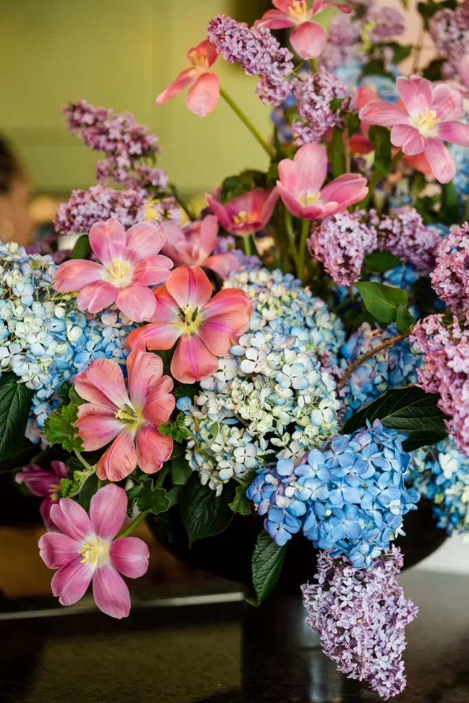 A vibrant bouquet of mixed flowers, featuring light pink tulips and clusters of blue and white hydrangeas, interspersed with purple lilacs. The lush display was the centerpiece at a Palo Alto Bat Mitzvah, set against a blurred green background.