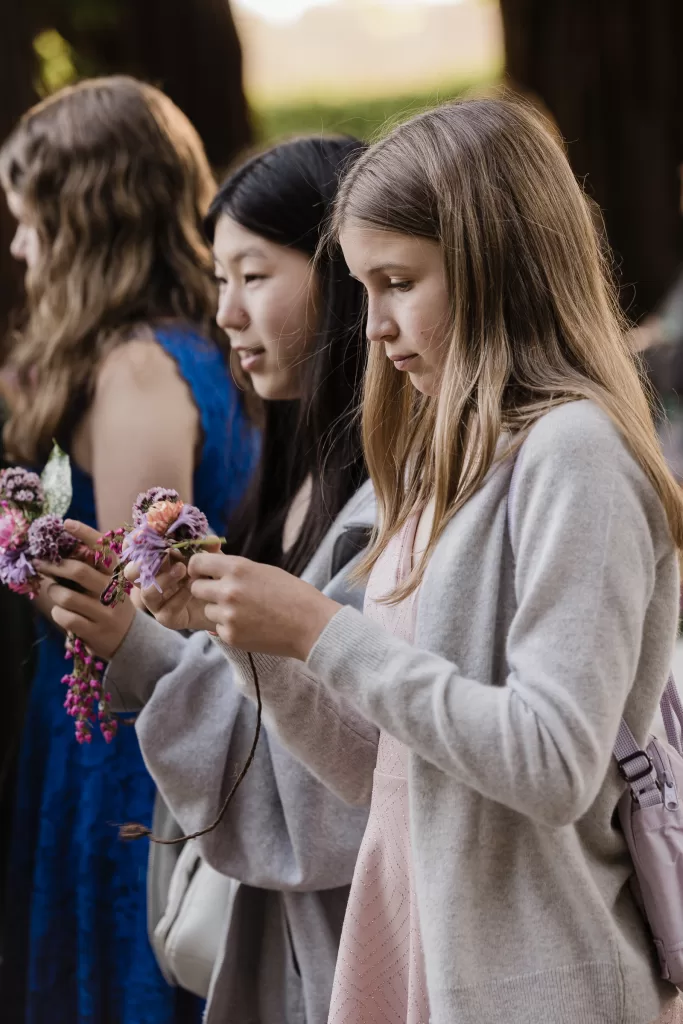 Three girls are standing in line, each holding colorful flower crowns or decorations. They seem focused on the items in their hands. All are dressed casually, with light jackets or cardigans. The blurred nature background suggests an outdoor Palo Alto Bat Mitzvah celebration.