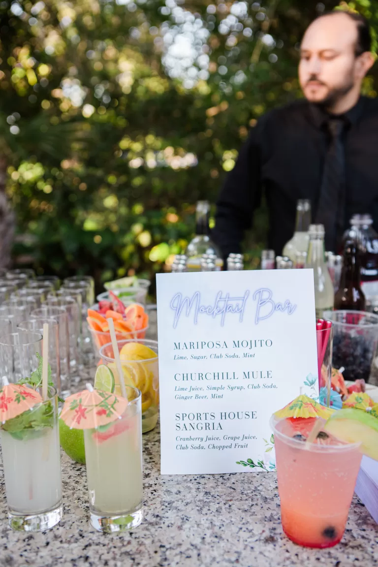 A bartender stands behind a mocktail bar set up outdoors at a Palo Alto Bat Mitzvah. On the bar counter are several colorful, garnished mocktails next to a menu that lists drinks including the Mariposa Mojito, Churchill Mule, and Sports House Sangria. Trees and greenery are visible in the background.