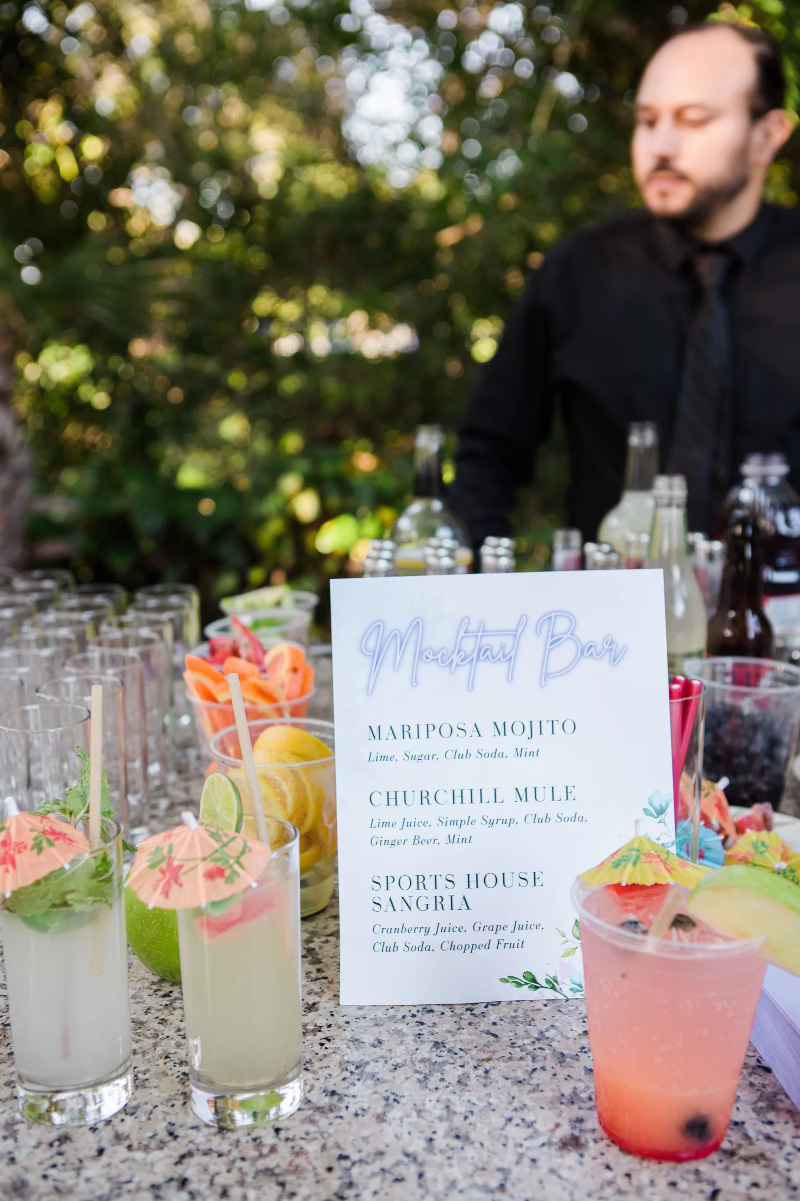 A bartender stands behind a mocktail bar set up outdoors at a Palo Alto Bat Mitzvah. On the bar counter are several colorful, garnished mocktails next to a menu that lists drinks including the Mariposa Mojito, Churchill Mule, and Sports House Sangria. Trees and greenery are visible in the background.
