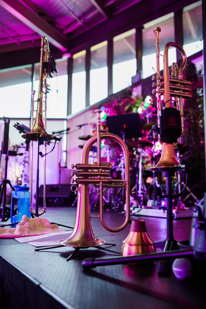 A music performance stage with various brass instruments, including trumpets and a trombone, placed on stands. A microphone lies in the foreground. Colorful lighting illuminates the background, reflecting off the instruments, creating a vibrant atmosphere perfect for a Palo Alto Bat Mitzvah.
