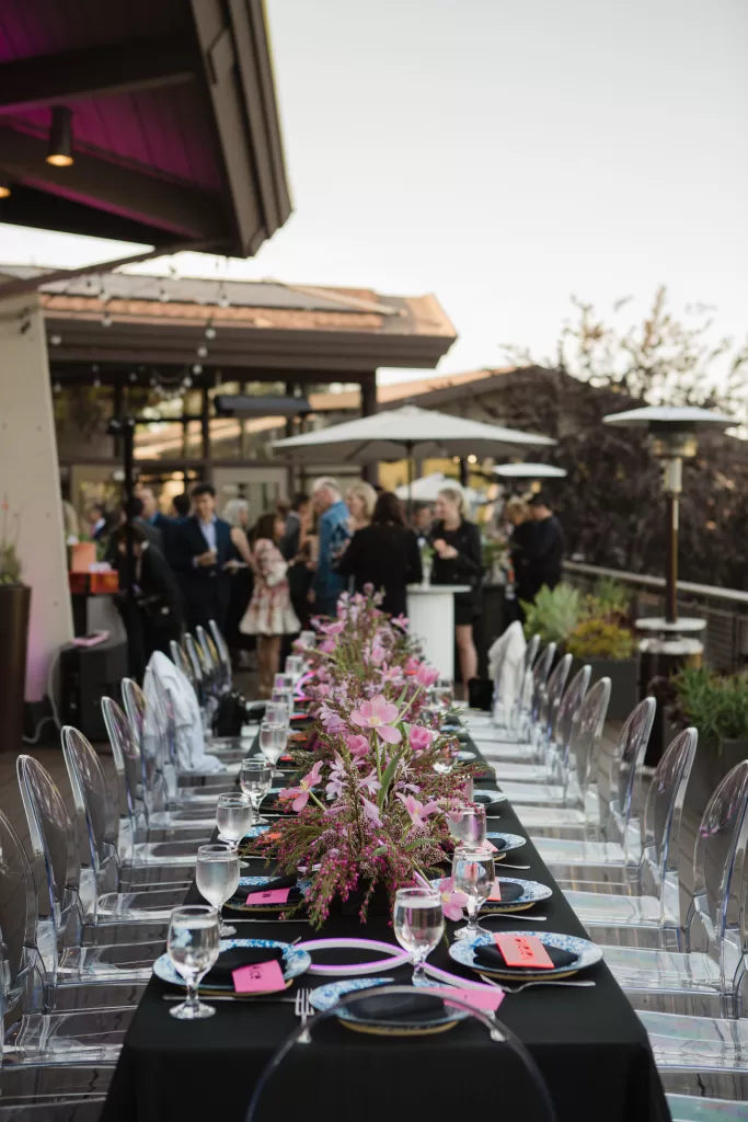 A long outdoor dining table with a black tablecloth, set with plates, silverware, and glasses, is adorned with pink floral centerpieces for the Palo Alto Bat Mitzvah. Transparent chairs surround the table. In the background, people are conversing near a building with string lights overhead.