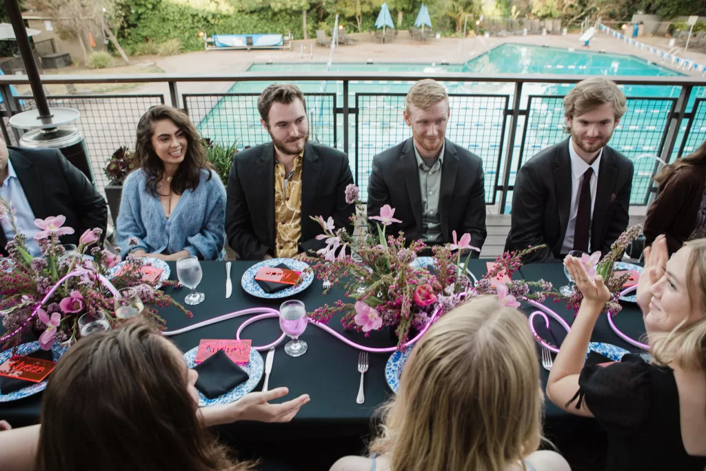 A group of people are seated around a rectangular, black-tableclothed table decorated with pink flowers and ribbons, set beside a pool at a Palo Alto Bat Mitzvah. Some are dressed formally in suits and dresses, engaged in conversation and appearing to enjoy each other's company.