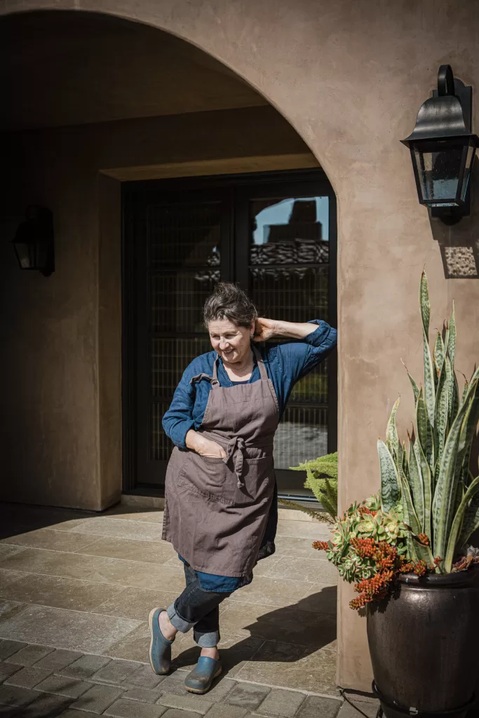 A person with short hair stands under an arched doorway, wearing a blue shirt, brown apron, and blue clogs. They lean against the wall with one hand behind their neck, next to a pot with green and red plants. The background includes a brown wall and a black lantern.