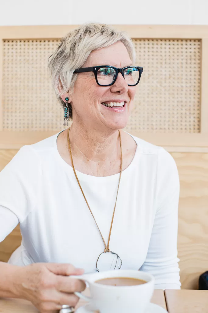 A smiling kelly zjilstra with short, blonde hair and black-framed glasses sits at a table holding a cup of coffee. She is wearing a white shirt, drop earrings, and a long necklace with a circular pendant. The background features a wooden panel with a woven design.