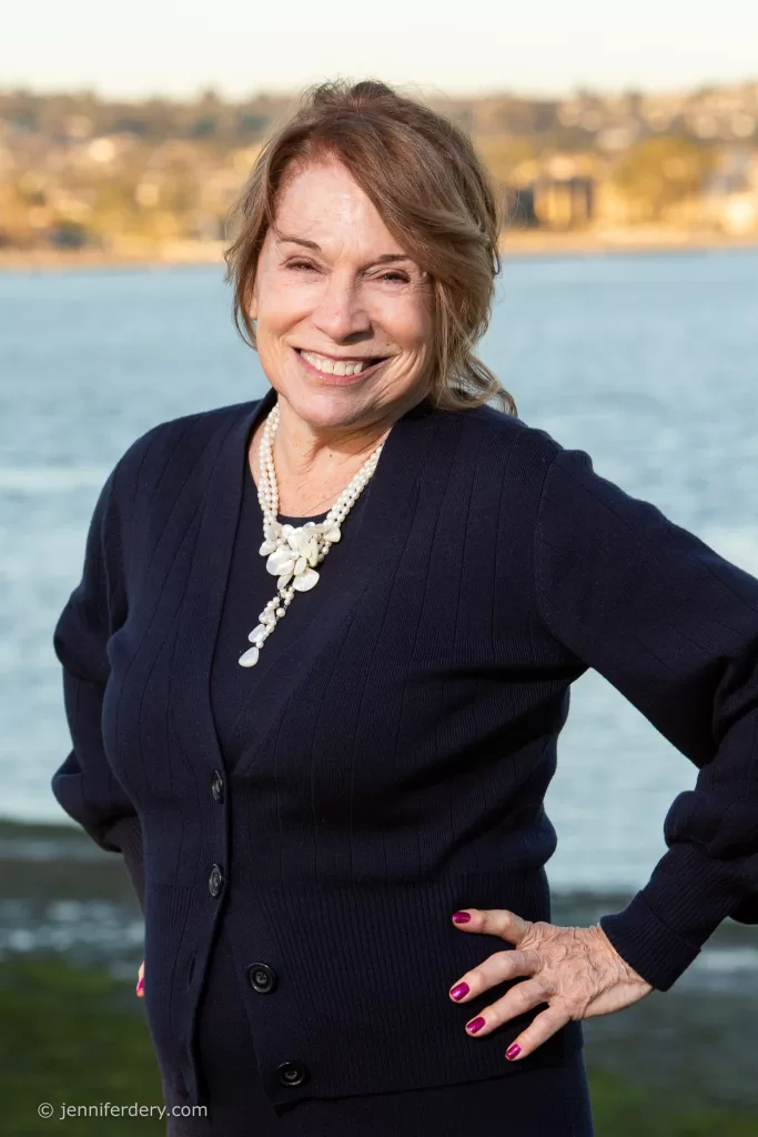 A woman with shoulder-length light brown hair, wearing a navy cardigan and a statement necklace, smiles at the camera. She has one hand on her hip, standing outdoors near a body of water with a blurred landscape in the background.