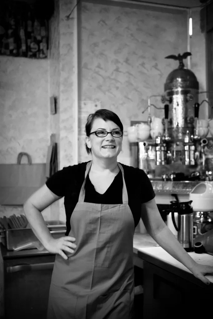 A woman wearing glasses and a light-colored apron stands smiling in a kitchen. She has one hand on her hip and is leaning against a counter. Behind her, a coffee machine and other kitchen appliances are visible. The image is in black and white.