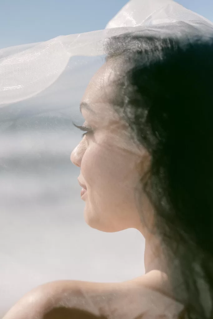 A close-up side profile of a person with long dark hair, partially covered by a delicate sheer fabric. The background is blurred but suggests a coastal setting with a bright sky, capturing the essence of wedding photography. The person's expression is serene and peaceful.
