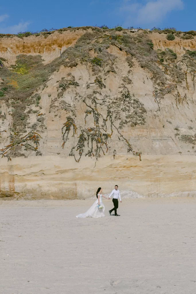 A couple, dressed in wedding attire, walks hand in hand on a sandy beach. The bride wears a flowing white gown, holding a bouquet, and the groom wears a white shirt and black pants. Behind them, a large cliff with sparse vegetation rises against a blue sky—perfect for stunning wedding photography.