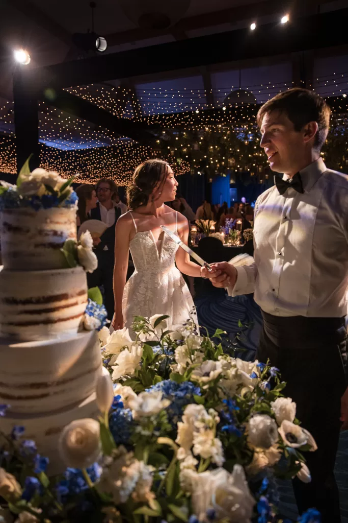 At their Coronado wedding, the bride and groom stand together, ready to cut a multi-tiered cake adorned with white and blue flowers. The venue is elegantly lit with string lights, casting a warm and festive glow over the celebration.
