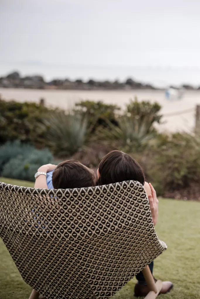 couple snuggling in a chair in front of the beach at the hotel del coronado in san diego. white dress and blue button-down with navy pants