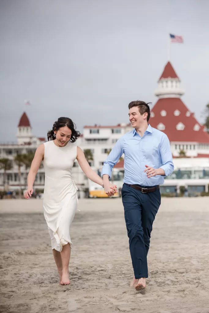 A couple joyfully runs barefoot on the beach, holding hands. The woman is wearing a sleeveless white dress, and the man is dressed in a light blue shirt and navy pants. A historic building with red turrets and an American flag in the background completes this perfect hotel del Coronado engagement session scene.