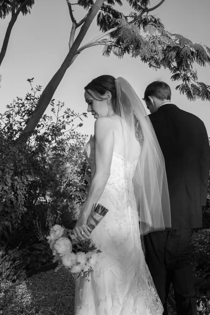A bride in a lace wedding dress and veil stands beside a groom in a dark suit, both facing a garden with lush greenery and tall trees. The bride holds a bouquet of flowers and the scene is captured in black and white.