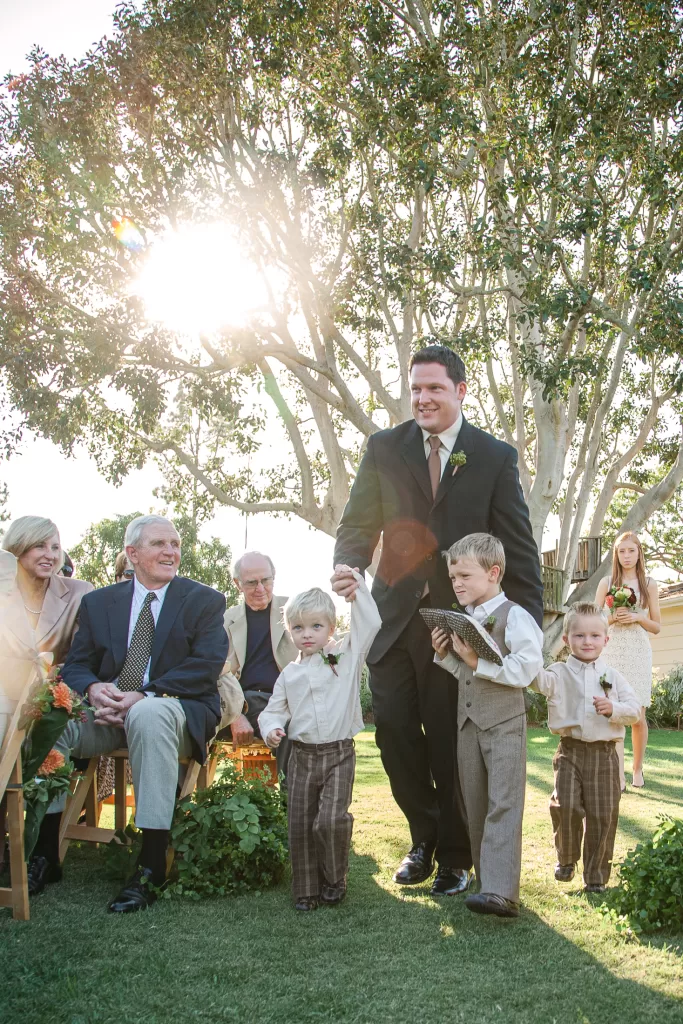 A man in a black suit escorts three young boys, holding hands with one of them, down an outdoor aisle. Sunlight filters through trees in the background. Seated guests, including older individuals, watch with smiles. A woman follows from behind, also observing with a smile.