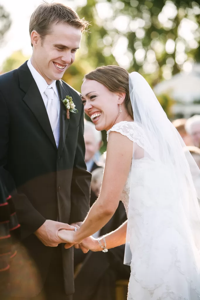 A bride in a white wedding gown and veil laughs heartily while holding hands with the groom, who is wearing a black suit and tie. They are standing outdoors with trees and blurred guests visible in the background.